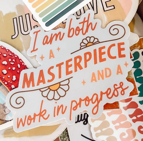 Image of a sticker that says "I am both a masterpiece and a work in progress" with orange writing and daisy flowers