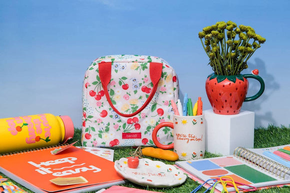 Colorful stationery items including a floral bag, mug, and notebook on grass with a blue sky.