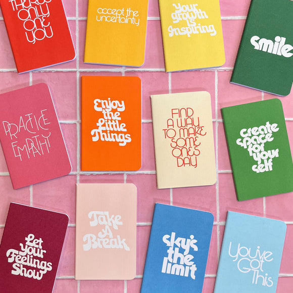 Each of the Ban-do Wellness Notebooks in the set laid out. There is a red one, a pink one, a maroon one, an orange one, a light pink one, two yellows, a light blue and dark blue, and a dark green and light green. Each say a different inspirational quote.