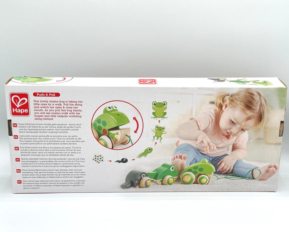 Back of the Hape Frog Family Pull Along. It is white with a description of the product in red writing, an image of a little girl playing with the product, and a circle with a large light green frog and a red arrow demonstrating its mouth goes up and down. The product has a black tadpole with wooden wheels, a small dark green frog with wooden wheels, and a large light green frog with wooden wheels.