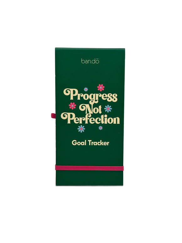 The Ban-do Progress Not Perfection goal tracker. It has a green cover that says, "Progress not perfection goal tracker" in cream writing with pink and blue flowers. There is a pink loop for a pen on the left side and a pink strap along the bottom to keep the notebook shut.