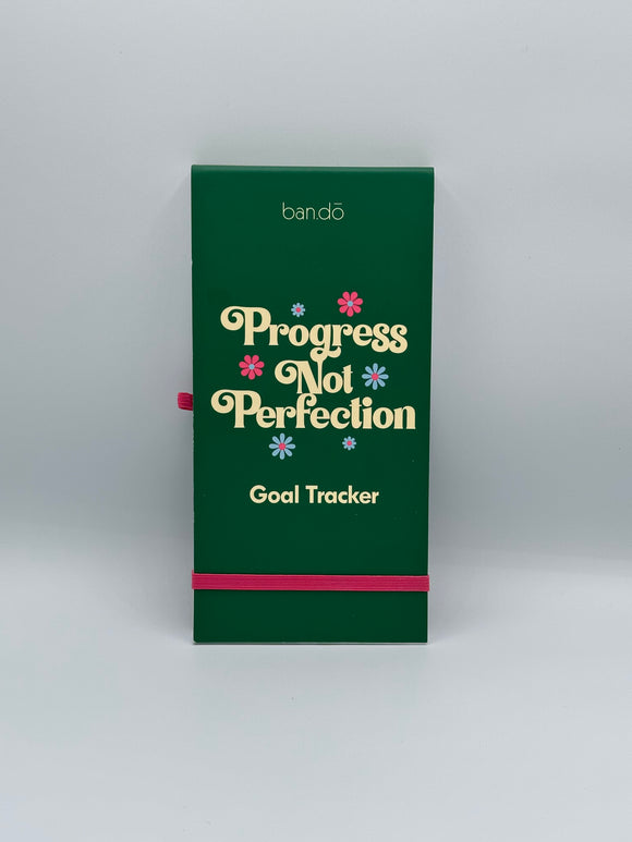 The Ban-do Progress Not Perfection goal tracker. It has a green cover that says, "Progress not perfection goal tracker" in cream writing with pink and blue flowers. There is a pink loop for a pen on the left side and a pink strap along the bottom to keep the notebook shut.