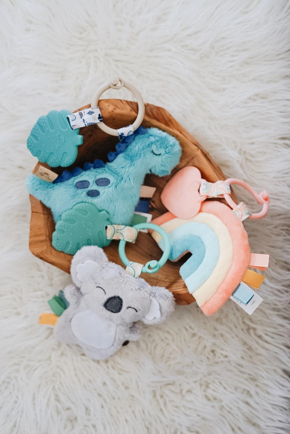 Koala, dinosaur, and rainbow Itzy Ritzy Itzy Pals in a wooden bowl. The dinosaur is a blue plush attached to a grey ring with a plastic blue leaf. The koala is a grey plush attached to a blue ring with a plastic green leaf, and the rainbow is a pink, yellow, light blue, and teal plush attached to a pink ring with a plastic pink heart.