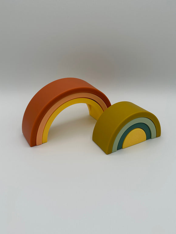 A silicone rainbow with 7 pieces that all nest within each other. On the right is the 3 outer pieces (from outside to inside) that are red, pink, and yellow. In front and to the right are the 3 inside pieces that are (from outside to inside) olive green, mint green, teal, and yellow. Each piece is an arc, except the yellow inner most piece is a semi-circle.