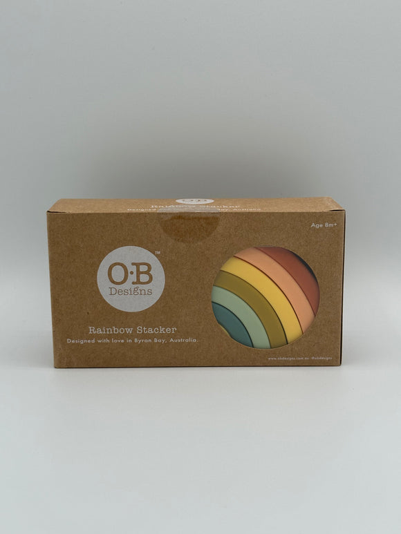 The silicone rainbow stacker in the package. The package is a cardboard box that says, "OB Designs, Rainbow Stacker, Designed with love in Byron Bay, Australia." On the right side is a clear circle cutout through which you can see the silicone stacker that is (from outside to inside) red, pink, yellow, olive green, mint green, and teal with a small visible piece of the yellow inside.
