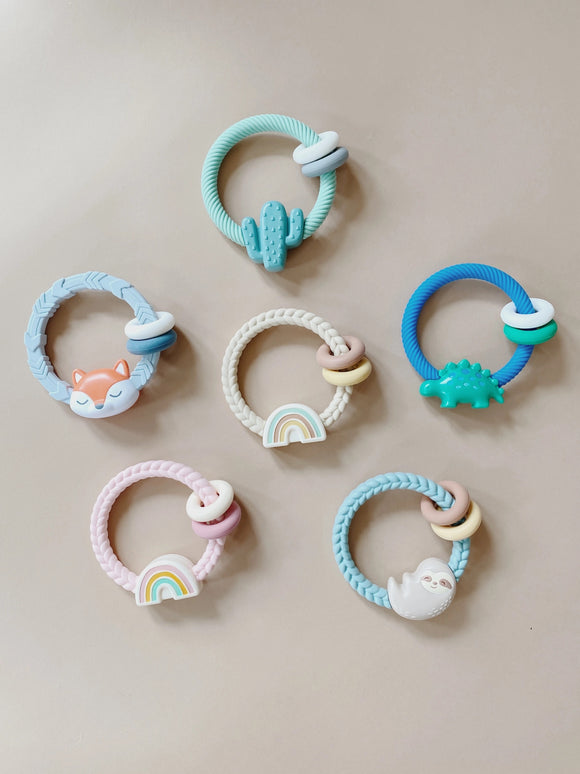 All of the Itzy Ritzy Ritzy Rattles in a circle formation. There is a teal ring with a teal cactus and smaller white and grey rings, a blue ring with an orange fox and smaller white and blue rings, a tan ring with a rainbow and smaller brown and yellow rings, a blue ring with a green dinosaur and smaller white and green rings, a pink ring with a rainbow and smaller tan and pink rings, and a blue ring with a grey sloth and smaller brown and yellow rings.