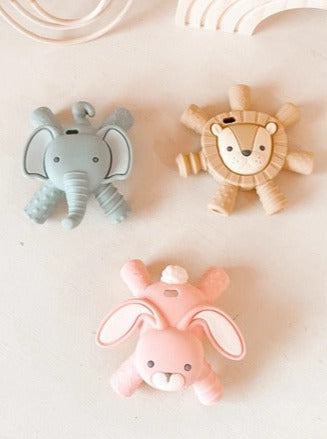 Grey elephant silicone Ritzy Teether with 4 legs coming off of each corner, tan lion silicone Ritzy Teether with 7 spikes coming off of its mane, and pink bunny silicone Ritzy Teether with 4 legs coming off of each corner.