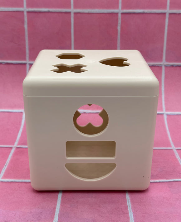 The empty Mushie Shape Sorting box. The box is white and shows the front of the box with the circle, rectangle, and semi-circle cutouts, and the top of the box with the hexagon, x, and heart shaped cutouts.