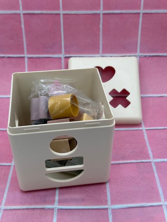 The Mushie Shape sorting box with the top taken off. In the background is the white top which has a heart, x, and hexagon cutout. In the front is the white box that shows the circle, rectangle, and semi-circle cutouts on the front. Inside the box are the 3D shapes which correspond to the shapes on the side of the box and are light purple, yellow, pink, sage green, and tan.