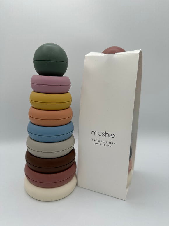 The rustic Mushie Stacking Rings tower out of the package next to the packaging. The tower contains 8 rings of descending order going up and a ball on top. From bottom to top, it has a white, dusty pink, brown, light grey, light blue, light orange, yellow, and light pink ring with a sage green ball. To the right is the packaging, which is a white rectangular paper wrapper. In thin, black font, it says, "mushie, stacking rings, 6 months - 3 years."