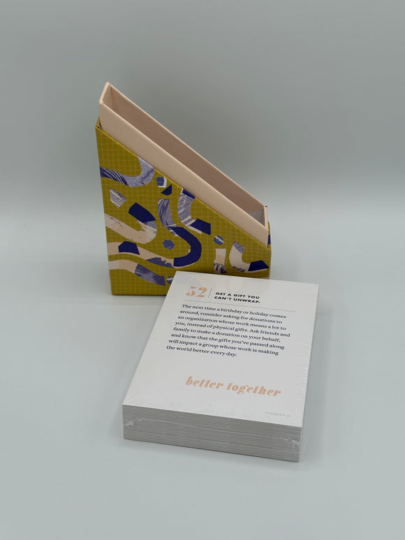 In the back is the Better Together box open. the light pink box is slanted and is sitting inside of the slanted chartreuse lid with dark blue and light pink squiggles. In the front is the stack of cards. Each card has a light pink number in the corner with a prompt written in dark blue and they all say, "better together" in light pink across the bottom.