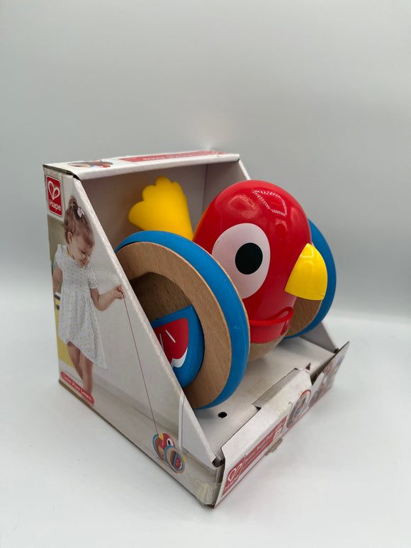 Hape Baby Bird Pull Along. It is a red bird with big eyes, a yellow beak and tail, and big wooden wheels that have blue rims pictured inside of a white box with an image of a little girl pulling the bird with a red string attached to it.