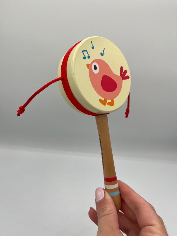 Hand holding the Hape Twittering Bird Rattle. The rattle is on a wooden stick with a light yellow drum on top with a pink bird with a red wing and 3 blue music notes on it. On the sides of the drum are two red strings.