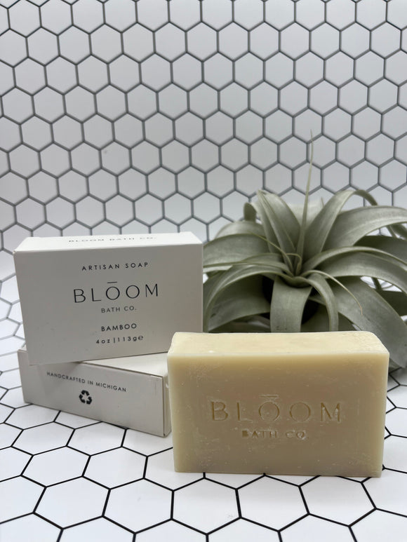 The bamboo Bloom body bar. The soap is in the front and says, "Bloom Bath Co", and the box is in the back stacked on top of a laying down box. It is white and says "Bloom Bath Co Bamboo". There is an air plant in the back corner.