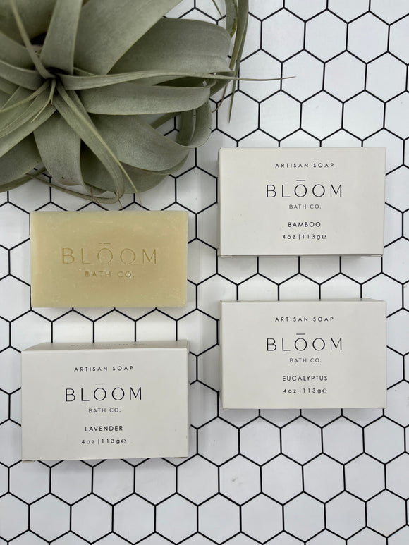 4 Bloom bath bars. One is out of the package and 3 are in the package. The bar is tan and says, "Bloom Bath Co", and the boxes are white and say, "Bloom Bath Co" with the scent of the soap (bamboo, lavender, eucalyptus).