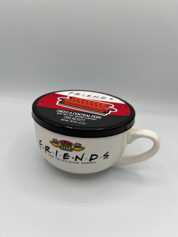 White mug shaped container that says, "friends, The Television Series" in black. Above the writing, there is an image of two yellow steaming coffee mugs outside of a green, yellow, and red sign that says, "central perk". The lid has one white stripe that says, "friends, the television series" in black, a red stripe with a red couch, and a black stripe that says, "I need a central perk, body butter - beurre corporel, Mad Beauty, 300 mL/10.1 fl oz."