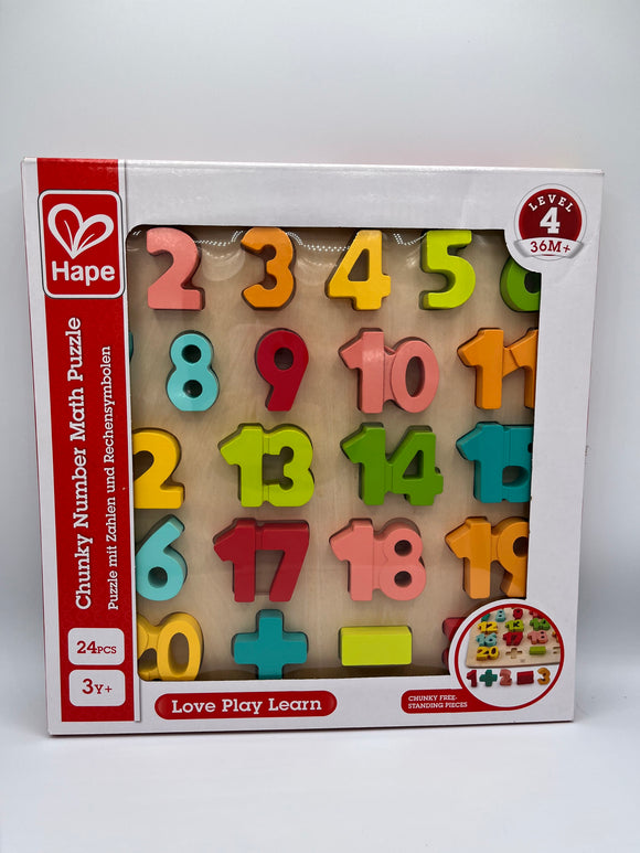 Hape Chunky Number Math Puzzle inside of the box. The box is white with a vertical red strip along the left side that says, "Chunky Number Math Puzzle, 27 pcs, 3y+". Along the bottom there is a red box that reads, "Love Play Learn". There is a clear film that showcases the product inside. It is a wooden board with numbers going up to 20 with a plus sign, minus sign, and equal sign in alternating colors of red, pink, orange, yellow, light green, dark green, dark teal, and light teal.