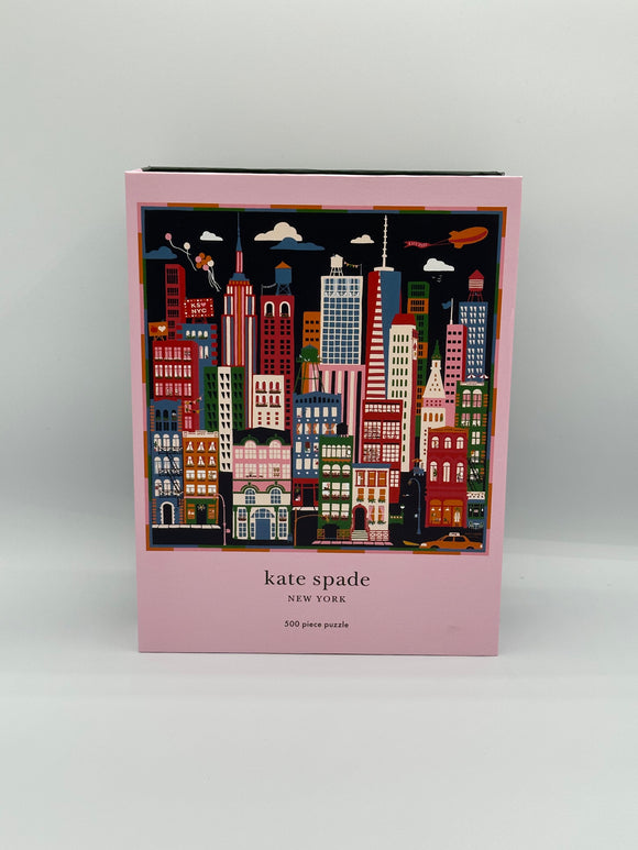 Light pink box with the image of the puzzle in the middle. The puzzle has a navy blue sky with a cityscape that has red, blue, pink, green, and orange skyscrapers. Underneath, it says, "kate spade, New York, 500 piece puzzle".