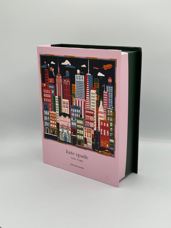 Sideview of the puzzle box. The front of the box is light pink with a picture of the puzzle that has a navy blue sky with red, white, pink, green, orange, and blue skyscrapers. Underneath, it says, "kate spade, New York, 500 piece puzzle". The back half of the box is dark green.