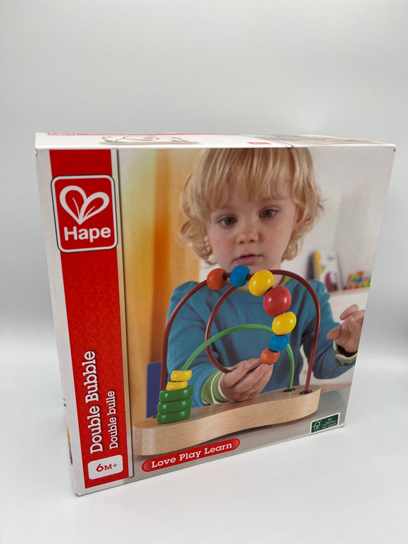 Hape Double Bubble box. It has a vertical red strip along the left side that reads, "Double Bubble, 6M+". The rest of the box contains an image of a blonde child wearing a blue shirt playing with the product. The product is a wooden platform with a green wire going from one corner to the opposite corner and a red loopy wire going from the other corners. The green wire has 3 green and two yellow rings, and the red has a 7 balls that are orange, blue, yellow, red, yellow, blue, and orange.
