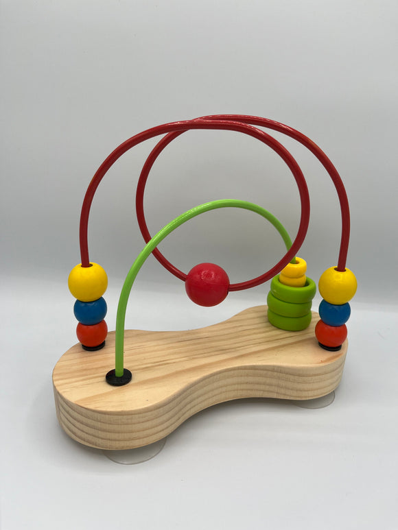 Hape Double Bubble outside of the box. It is a wooden peanut shaped platform with a light green wire going from one corner to the opposite corner and a red loopy wire going from the two other corners. The green wire has 3 green rings and 2 smaller yellow rings. The red wire has a red ball, a blue ball, and a yellow ball on each end and a red ball in the middle.