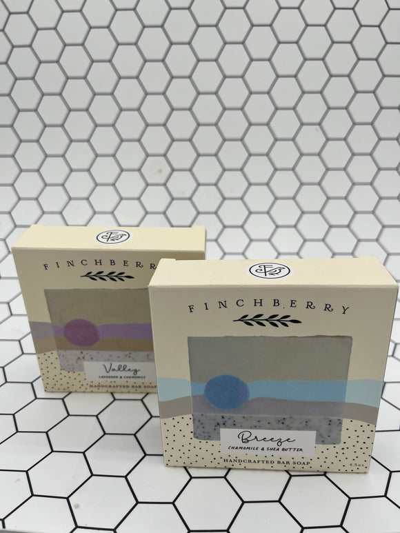 Both scents of the Finchberry Earth Soaps. Each are in tan boxes that say, "Finchberry" in black writing across the top and have a picture of the soap with the scent underneath. In front is the "Breeze" scent which is a grey soap with a blue stripe through the middle. In the back is the Valley scent which is a tan soap with a purple stripe through the middle.