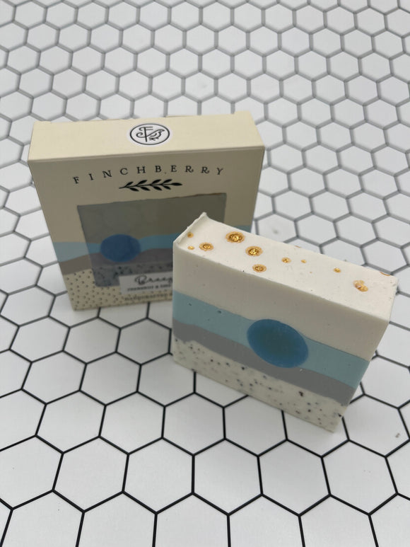 The soap and the box for the Fincheberry Breeze soap. In the back is the tan box it comes in that says, "Fincheberry" in black writing across the top with a photo of the soap. In the front is the soap. It is a layered soap with white with black speckles on the bottom, then grey, then light blue with a darker blue circle, then white, with gold flecks on top.