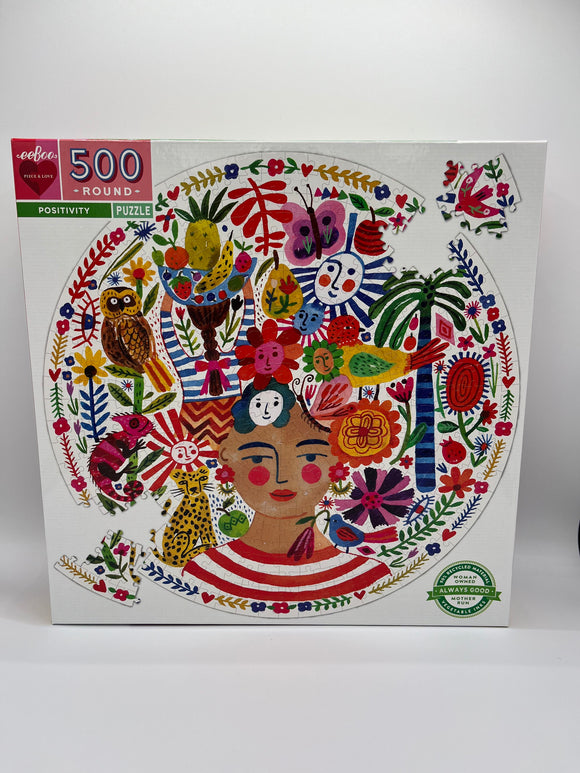 The front of the Eeboo Positivity Round Puzzle box. The box is white with a pink box in the corner that says, "500 Round" and a small green box underneath the pink box that says, "Positivity Puzzle". In the middle of the box it shows the puzzle. It is a woman, and her hair is made up of various flowers, fruits, plants, and shapes.