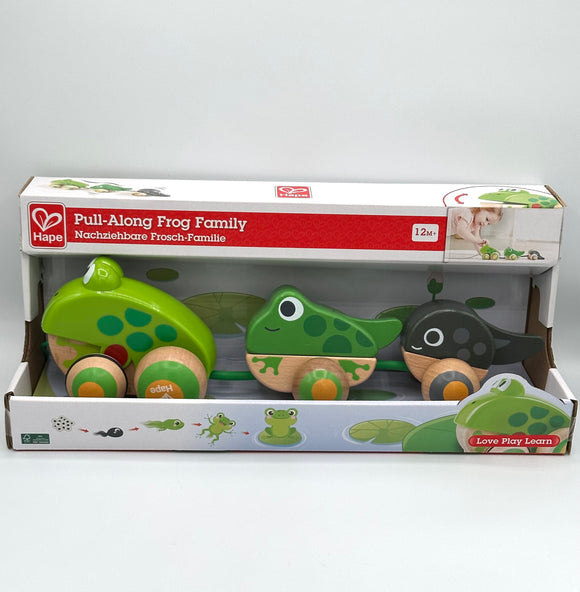 Hape Frog Family Pull-Along box. It's white with a red strip across the top that says, "Pull-Along Frog Family, 12M+". The middle the box showcases the product. There is a large light green frog with dark green spots and 4 wooden wheels with green and yellow concentric circles, a small dark green frog with darker green spots and two wooden wheels with green and yellow circles, and a black tadpole with grey spots and two wooden wheels with grey and yellow circles connected with a green rope.