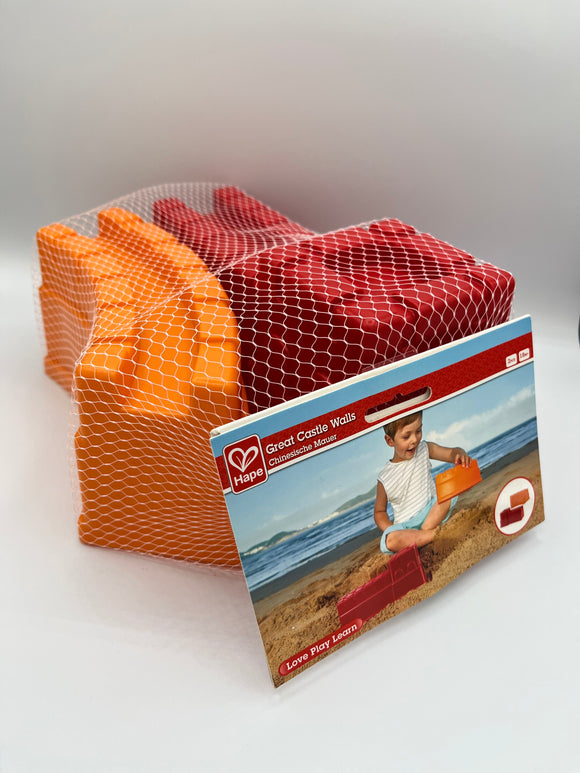 Hape Great Walls sand toys in a white mesh bag with a tag that showcases an image of a boy on the beach playing with the sand toys. One is a curved orange wall and the other is a straight red wall with a raised square tower.
