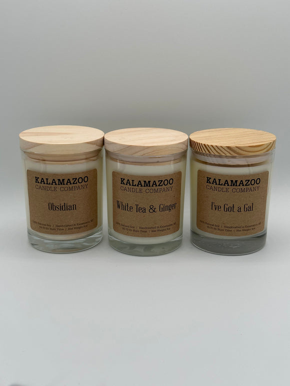 Each of the three scents of the Kalamazoo Candle Jars lined up. Each jar is clear with a white wood lid and a brown paper label that says, "Kalamazoo Candle Company" and the scent of the candle. You can see the white candle through the clear jar. (From left to right) the scents are obsidian, white tea and ginger, and I've got a gal".