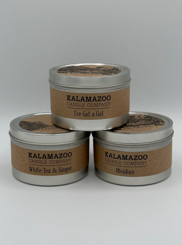 The three Kalamazoo Candle tins in a pyramid shape. Each tin is a small, metal, cylindrical tin that has a brown paper label that says, "Kalamazoo Candle Company" with the scent of the candle. On top is I've Got a Gal, on the bottom left is White Tea and Ginger, and on the bottom right is Obsidian.