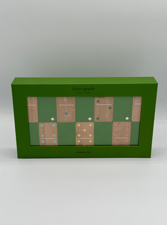 Green box that says, "kate spade, New York" on the top with a clear opening that shows two rows of alternating green and wood dominoes. Some of the dominoes have dark blue spots, some dark green spots, purple spots, light blue spots, yellow spots, and pink spots. The bottom of the box says, "Dominoes Set."