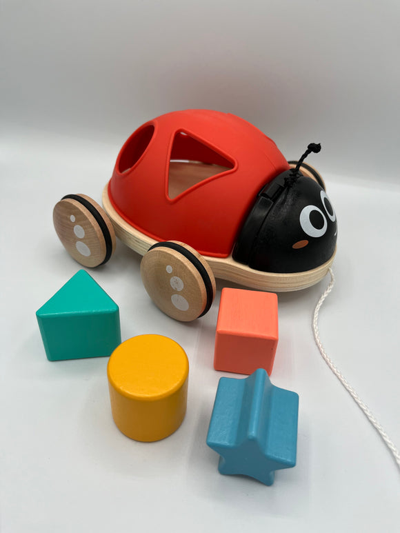 Hape Shape Sorter Ladybug. There is a red ladybug with cut outs on its back of a triangle, circle, square, and star. It has a black head with two rope antennas and 4 wooden wheels and a white string to pull it. In front there is a 3d green triangle, orange square, yellow circle, and blue star.