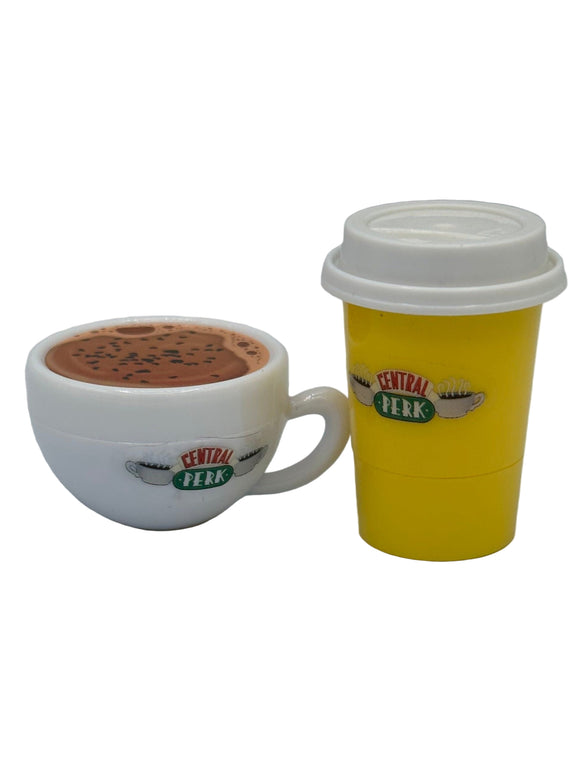 A white coffee mug shaped lip balm with brown coffee on top and on the front it says, "central perk" in red and green surrounded by two white steaming coffee mugs. On the right there is a yellow to-go coffee cup shaped lip balm with a white lid. It also says, "central perk" in red and green surrounded by two white steaming coffee mugs.