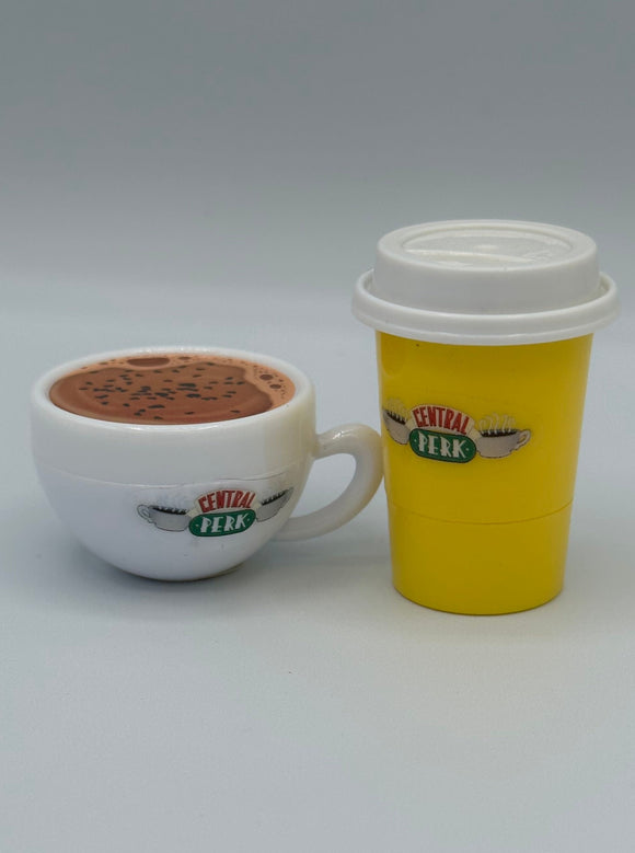 A white coffee mug shaped lip balm with brown coffee on top and on the front it says, "central perk" in red and green surrounded by two white steaming coffee mugs. On the right there is a yellow to-go coffee cup shaped lip balm with a white lid. It also says, "central perk" in red and green surrounded by two white steaming coffee mugs.
