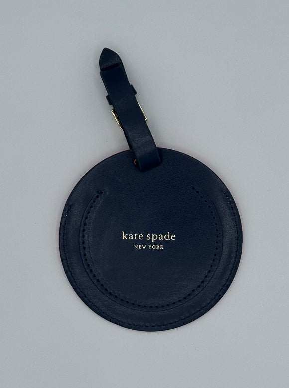 The back of the luggage tag. It is navy blue leather and has, "kate spade, New York" embossed in gold. At the top there is a navy blue loop with a gold buckle.