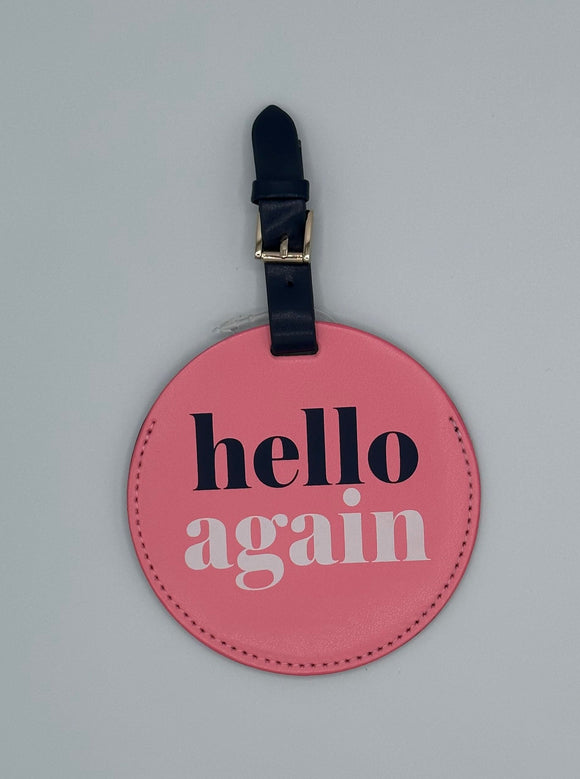 Pink, leather, circular luggage tag with a navy blue loop with a gold buckle. The tag says, "hello again" with hello in navy and again in white.