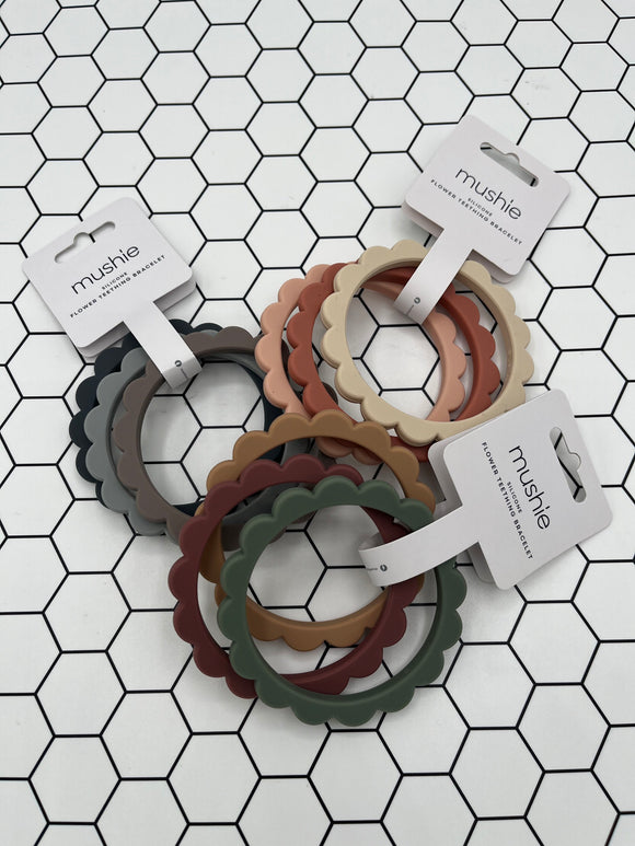 3 packs of the Mushie Flower Teething Bracelets. Each pack has 3 flower shaped rings. The pack in the back left has a navy, grey, and taupe ring. The pack in the back right has a light pink, rust, and tan ring. The pack in the front has a brown, maroon, and sage green ring.
