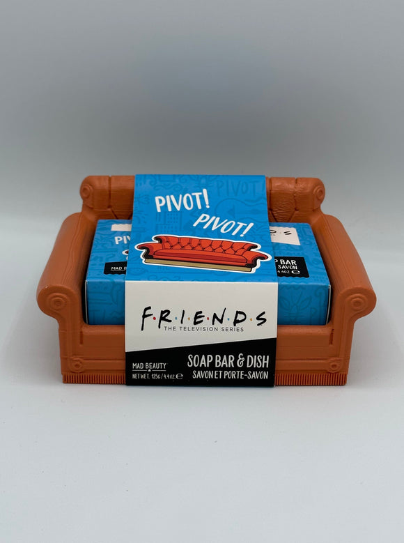 Brown, plastic couch soap dish. Sitting inside the couch is a bar of soap in a blue box. Around the couch is a paper label. The top half is blue and says, "pivot! pivot!" in white writing with a photo of a brown couch. The bottom half is half white and half black. The white half says, "Friends, the television series" in black writing, and the black half says, "Mad Beauty, Net Wet. 125 g/4.4 oz, Soap bar & dish, Savon et porte-savon."