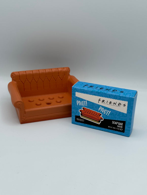 On the left is a brown, plastic couch shaped soap dish. On the left is a blue box. On the top of the box, it says, "friends, the television series" in black writing. On the front of the box, it says, "friends, the television series" in black writing on a white rectangle and, "Pivot! Pivot!" in white writing above an image of a brown couch. At the bottom of the front of the box there is a black rectangle that says, "soap bar, savon, net wt 125g/4.4 oz."