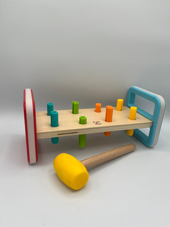 Hape Rainbow Pounder. It is a wooden plank of wood between two rectangles, one is red and one is light blue. There are 8 pegs going through the wood plank. 2 are light blue, 2 are light green, 2 are orange, and 2 are yellow. There is a wooden mallet with a yellow head.