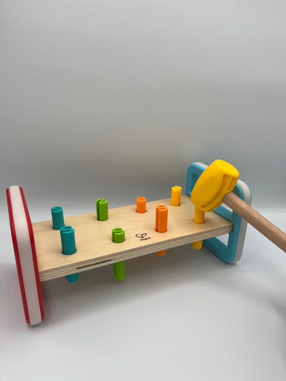 Hape Rainbow Pounder. It is a wooden plank of wood between two rectangles, one is red and one is light blue. There are 8 pegs going through the wood plank. 2 are light blue, 2 are light green, 2 are orange, and 2 are yellow. There is a wooden mallet with a yellow head pounding one of the yellow pegs.