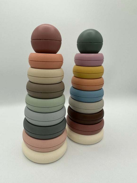The original (left) and rustic (right) Mushie Stacking Rings. Each tower contains 8 rings of descending sizes going up and a ball on top. The original tower (from bottom to top) has a white, light pink, sage green, light blue, mint green, taupe, cream, and peach ring with a dusty blush pink ball on top. The rustic tower (from bottom to top) has  a white, dusty pink, brown, light grey, light blue, light orange, yellow, and light pink ring with a sage green ball on top.