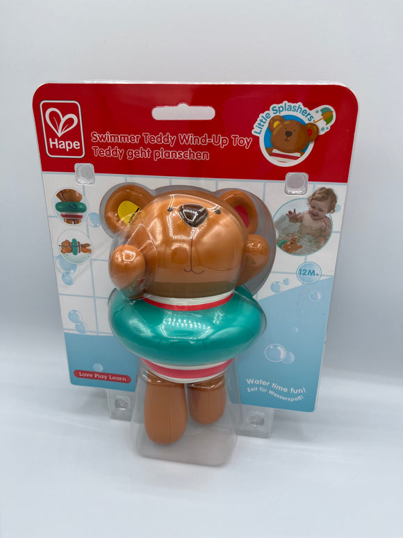Hape Swimmer Teddy in the package. Along the top is a red strip that says, "Swimmer Teddy Wind-Up Toy, Teddy geht planschen". Behind the teddy is a bathtub scene with white tile and blue water and bubbles. In the top right corner is an image of a baby playing with the teddy in the bath. The teddy is brown with a red and white stripe bathing suit and a teal inner tube around it.