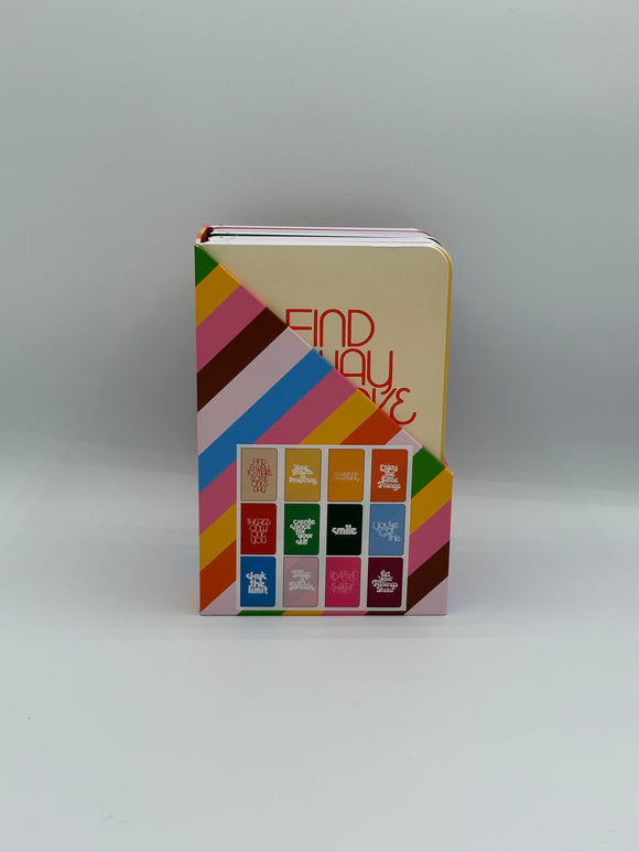 The Ban-do Wellness Notebook set. Shows them in the box they come in which has green, yellow, pink, brown, blue, and red diagonal stripes on it with an image showing each of the 12 notebooks.