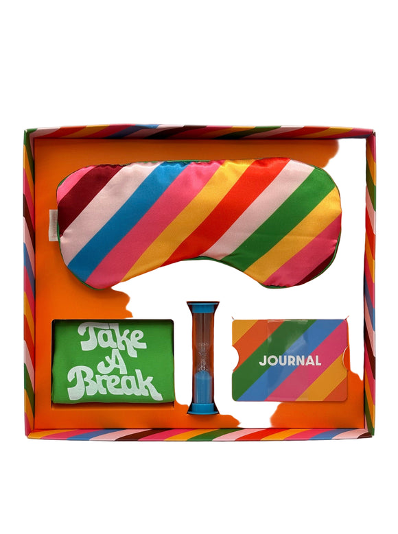 The inside of the Ban-do Unplug Kit box. The kit includes an eye mask with pink, brown, blue, yellow, red, and green diagonal stripes, a green phone sack that says, "Take a Break" in white writing, a small blue timer, and a small stack of journal prompt cards with yellow, orange, green, blue, and pink stripes and says 'journal' in white writing.