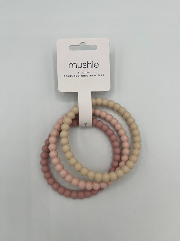 The Mushie Pearl Teething Bracelet Set in the package. From right to left, there is a dusty pink, light pink, and cream silicone ring all with a pearl texture. They are held together by a white paper package at the top that says, "mushie, silcone, pearl teething bracelet," in thin, black font.
