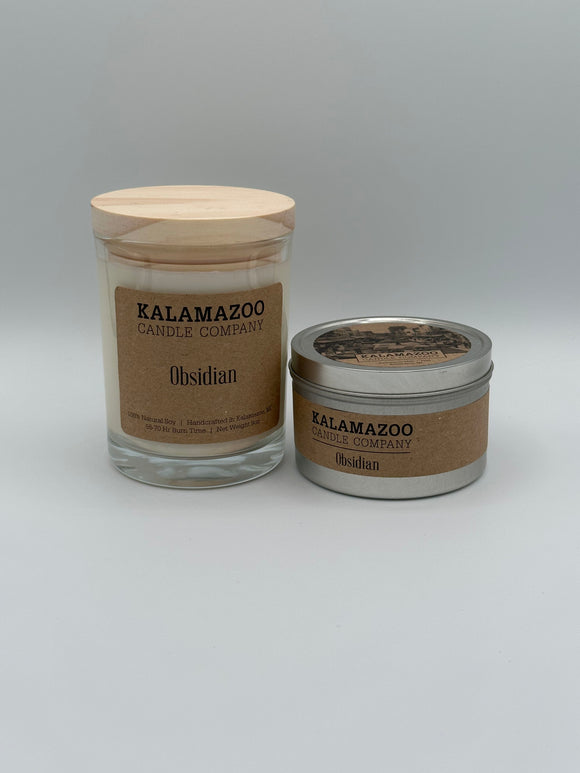The Obisidian Kalamazoo Candle jar next to the Obsidian Kalamazoo Candle tin. The jar is clear through which you can see the white candle with a light wood lid and a brown paper label that reads, "Kalamazoo Candle Company, Obsidian". The tin is a small cylindrical metal tin with a brown paper label that says, "Kalamazoo Candle Company, Obsidian". 