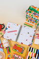Colorful stationery items including a planner, markers, and a lunch bag on a yellow surface.